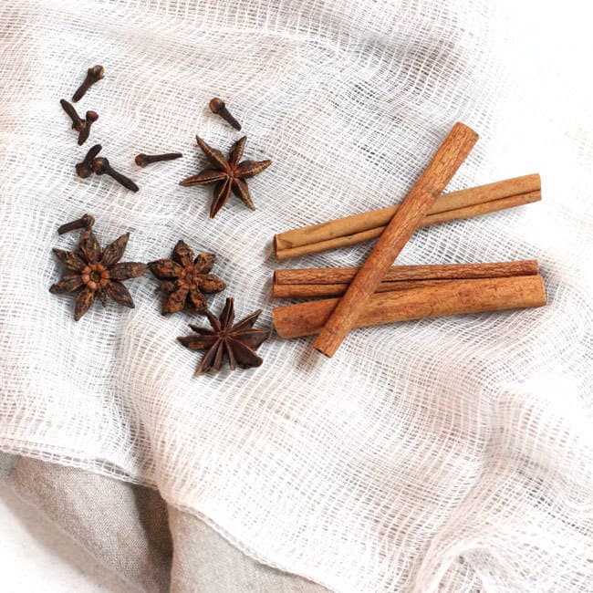 Overhead shot of cinnamon sticks, star anise, and whole cloves, on a white cheesecloth.