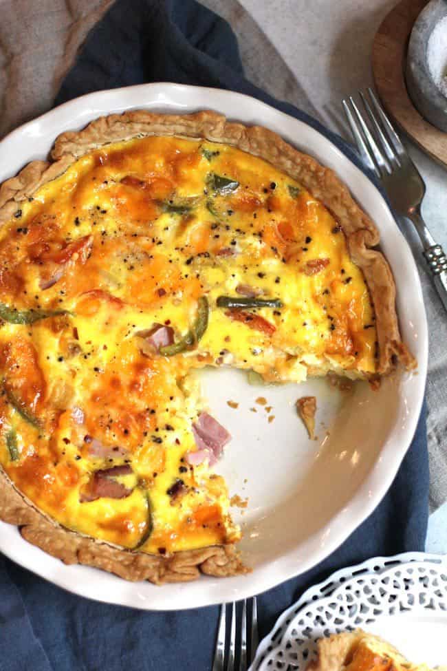 Overhead shot of a ham and cheese quiche in a white pie plate, with one slice missing, on a blue napkin.