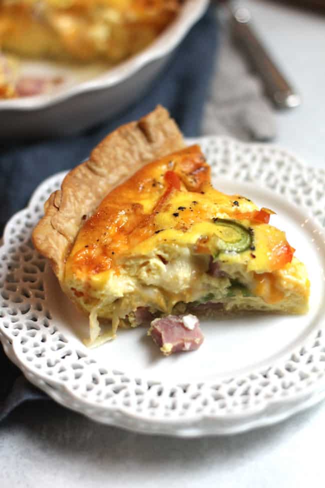 Side shot of a slice of ham and cheese quiche, on a white plate, with the quiche in the background.