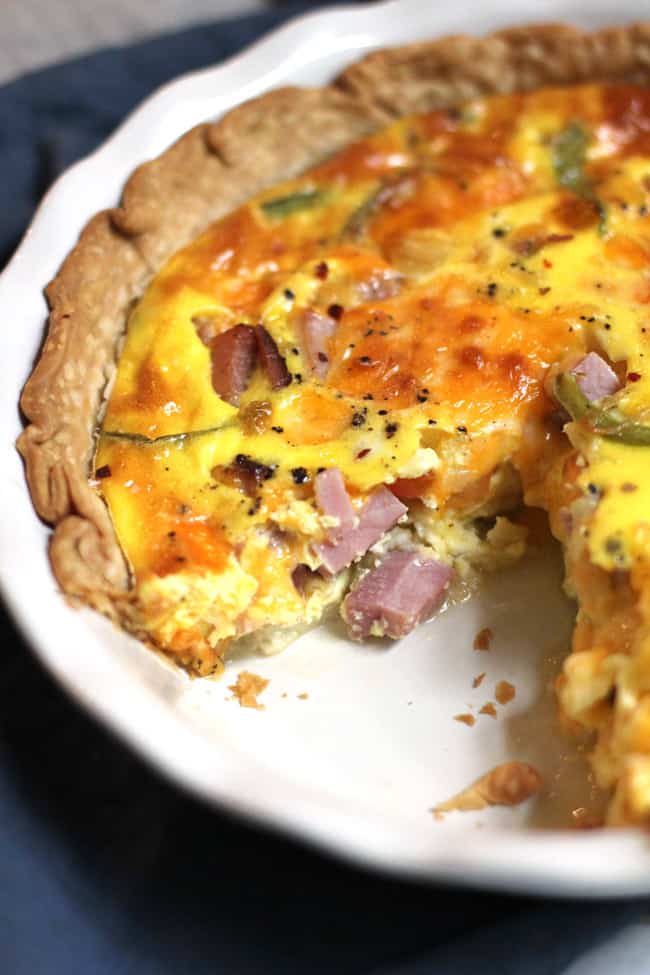 Overhead shot of a half of a ham and cheese quiche in a white pie plate, on a blue napkin.