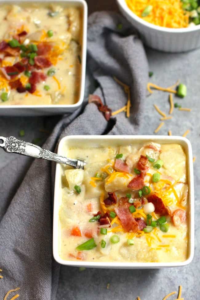 Overhead shot of two square white bowls of creamy potato soup, with toppings of bacon, cheese, and green onion, on a gray background.