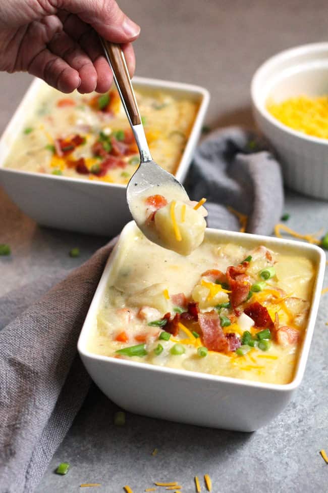 Side shot of two square white bowls of creamy potato soup, with toppings of bacon, cheese, and green onion, with my hand scooping out a spoonful, all on a gray background.