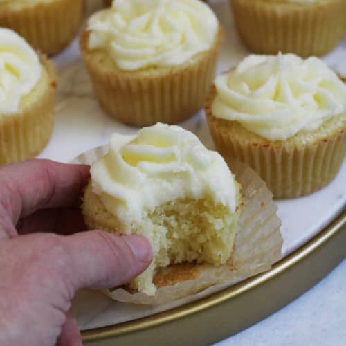 Side shot of my hand reaching for a lemon cupcake that has a bite out of it, on a white tray with other cupcakes.