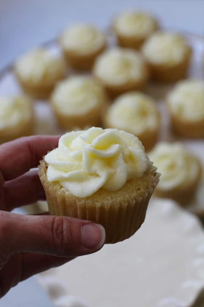 Side shot of my hand holding one lemon cupcake, with lemon cupcakes in the background.
