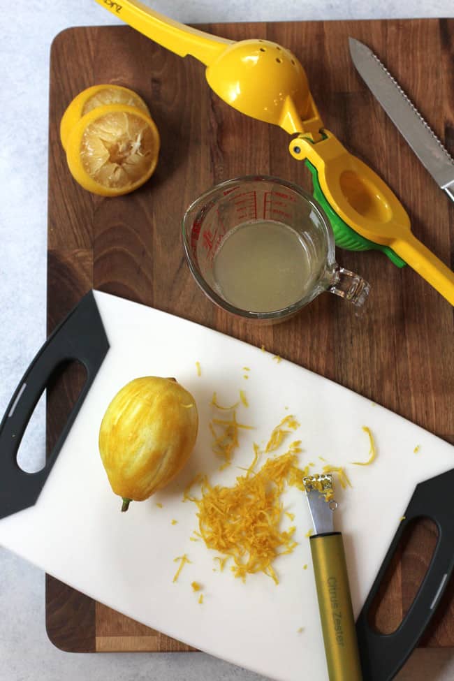 Overhead shot of a lemon juicer next to squeezed lemon juice, and a zester next to a lemon, all on a wooden background.