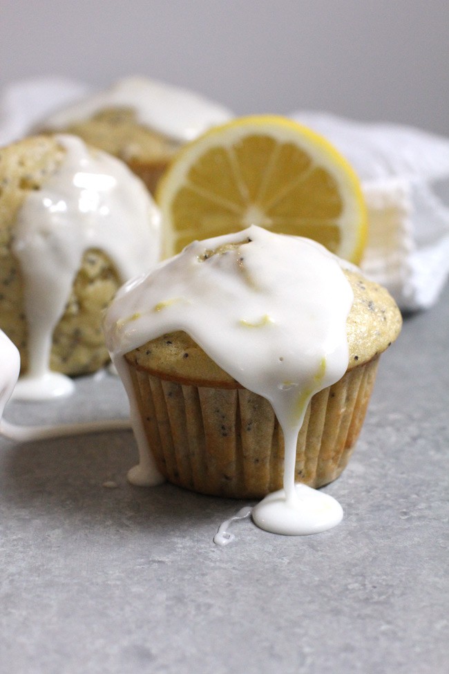 Side shot of glazed poppy lemon poppy seed muffins, with glaze dripping off, on a gray background with white napkin.