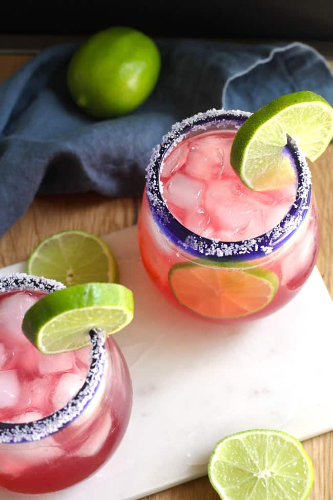 Overhead shot of two cranberry margaritas, in blue rimmed glasses, with a salt rim and lime wedge, on a white board with a blue napkin.