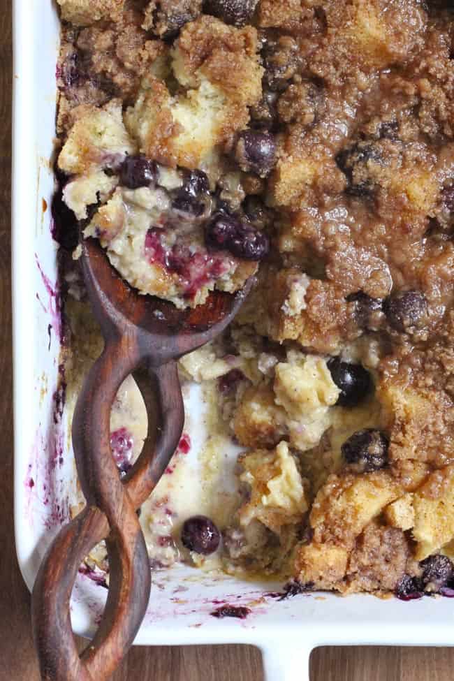 Overhead shot of one/fourth of a blueberry French toast casserole, with a serving removed, and a wooden spoon inside it.