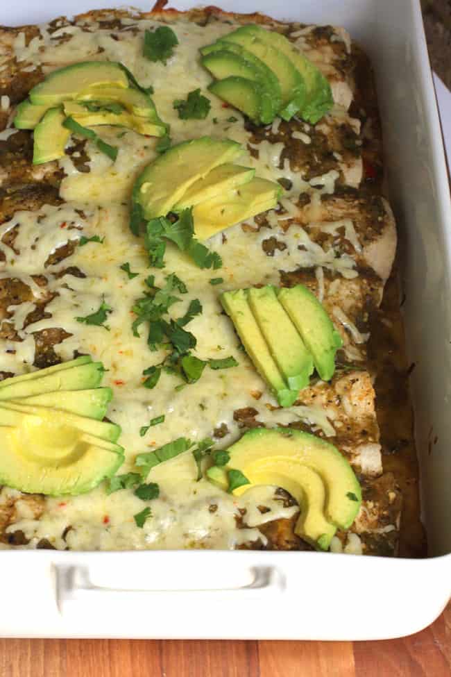 Side shot of Green Chile Chicken Enchiladas in a large white casserole dish, with avocado slices on top.