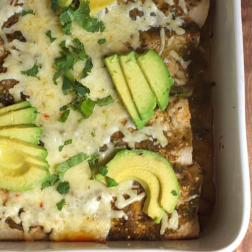 Overhead shot of part of a white casserole dish of Green Chile Chicken Enchiladas with avocado slices on top.
