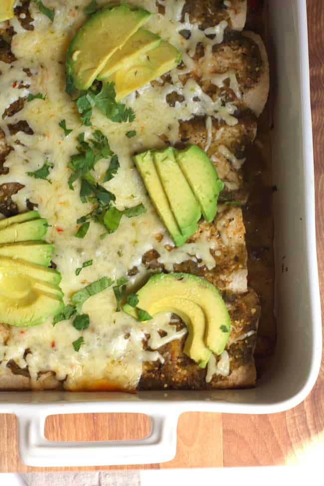 Overhead shot of part of a white casserole dish of Green Chile Chicken Enchiladas with avocado slices on top.