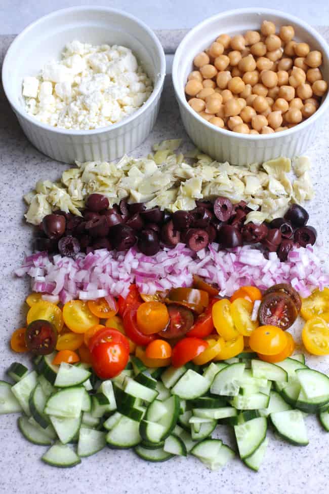 Overhead shot of the chopped ingredients in the pasta salad, lined up, with bowls of feta cheese and chickpeas.