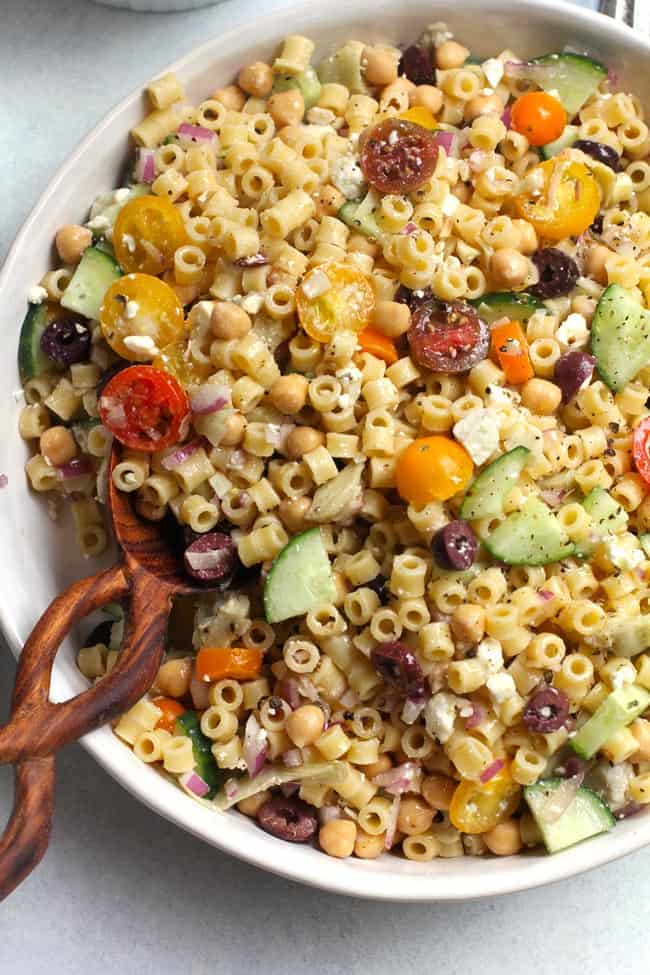 Overhead shot of a partial bowl of Chopped Greek Pasta Salad, with a wooden spoon, on a white background.