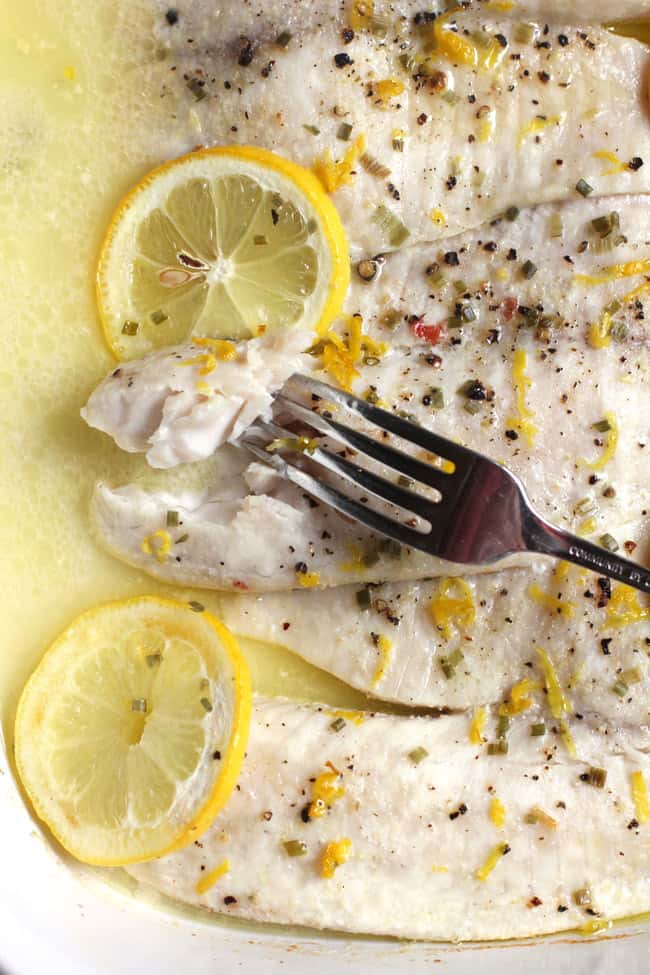 Close-up shot of Easy Baked Lemon Butter Tilapia, with lemon slices and sauce, and a fork stuck in a piece of fish.