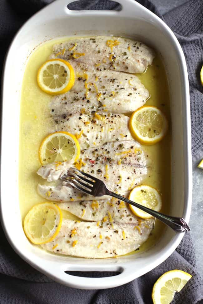 Overhead shot of a white casserole dish with four large pieces of lemon butter tilapia, with lemon slices on top.
