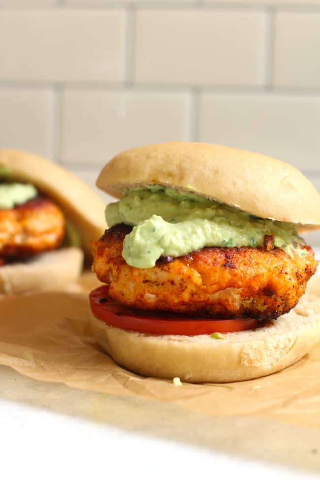 Side shot of a blackened salmon burger with Avocado Crema, on a wheat bun with a white background.