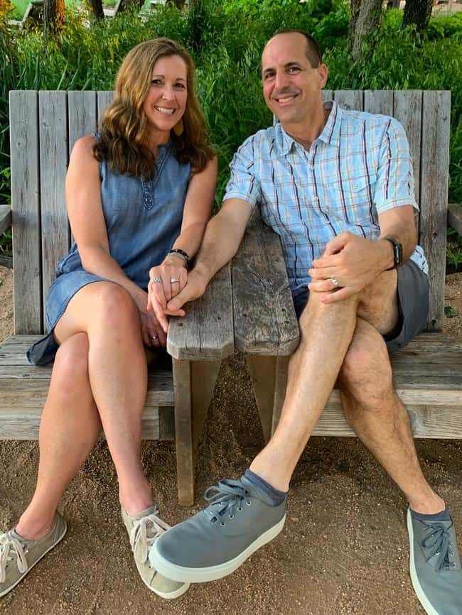 Sue and Mike, sitting on some outdoor chairs, holding hands.