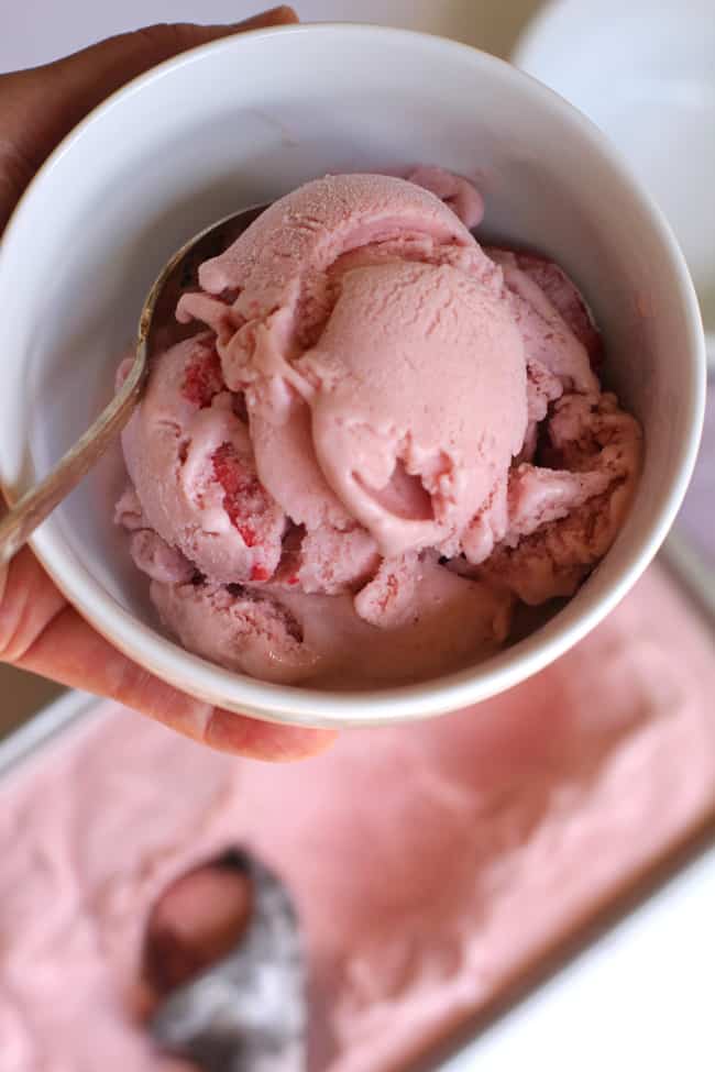 Overhead shot of my hand holding a round white bowl of strawberry ice cream, with a larger container in the background.