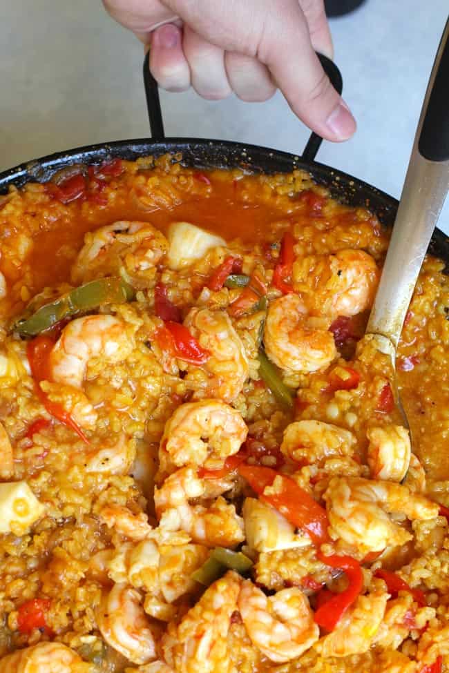 Overhead shot of Spanish Seafood Paella, in a large flat paella pan, with a hand lifting the pan.