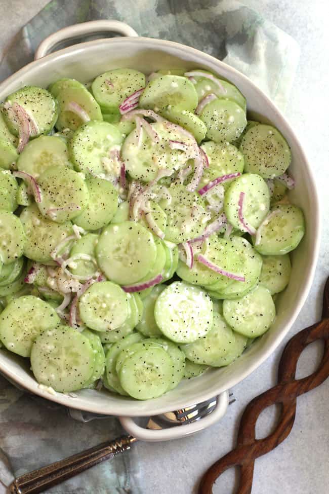 Overhead shot of creamy cucumber salad in a flat white bowl with handles, and a wooden spoon next to it.