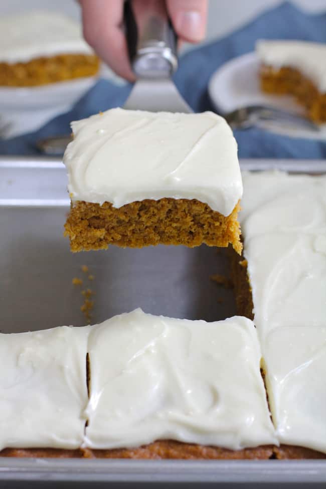 Side shot of a hand using a spatula to lift a piece of pumpkin cake from a pan, with a blue napkin in the background.