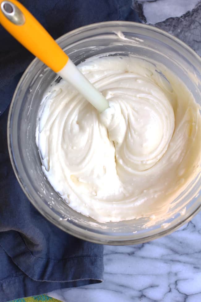 Overhead shot of a bowl of cream cheese frosting, with a rubber scraper inside, on a white and gray background.