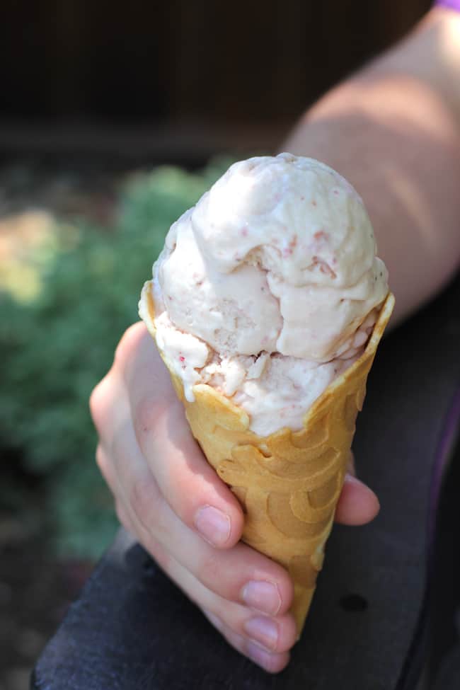 Side shot of a hand holding a strawberry ice cream cone, sitting outside on a deck chair.