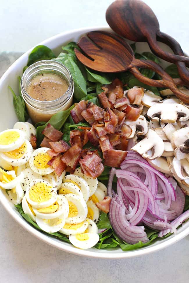 Overhead shot of a large white bowl of spinach salad ingredients in sections, with a jar of Honey Dijon dressing.