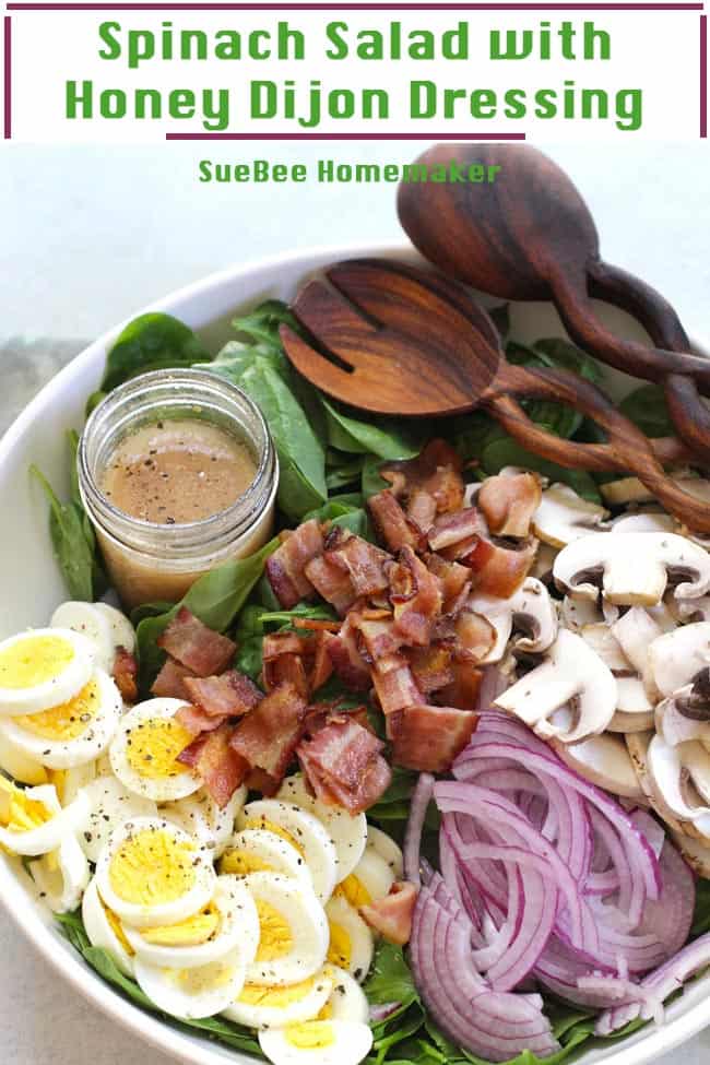 A large white bowl with spinach salad - eggs, bacon, mushrooms, red onions, Honey Dijon dressing, with wooden spoons.