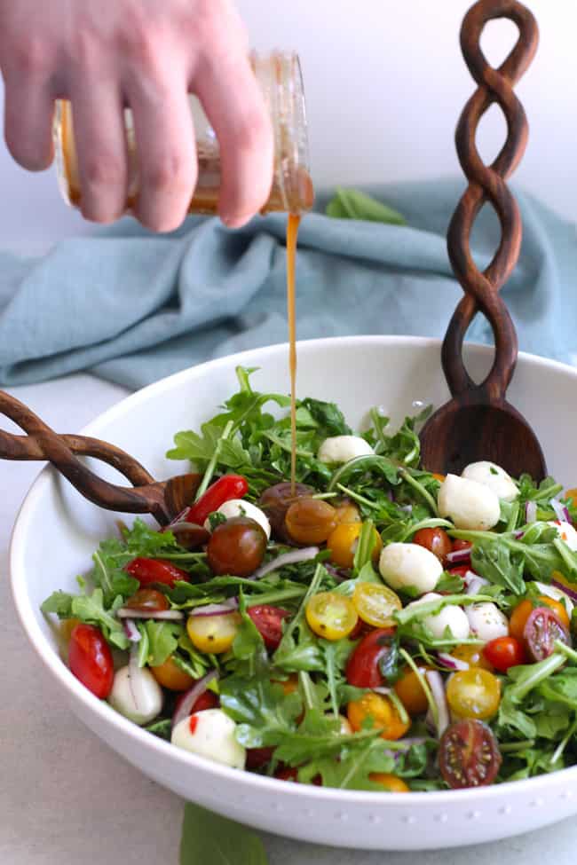 Side shot of a hand drizzling salad dressing on a bowl of arugula Caprese salad.