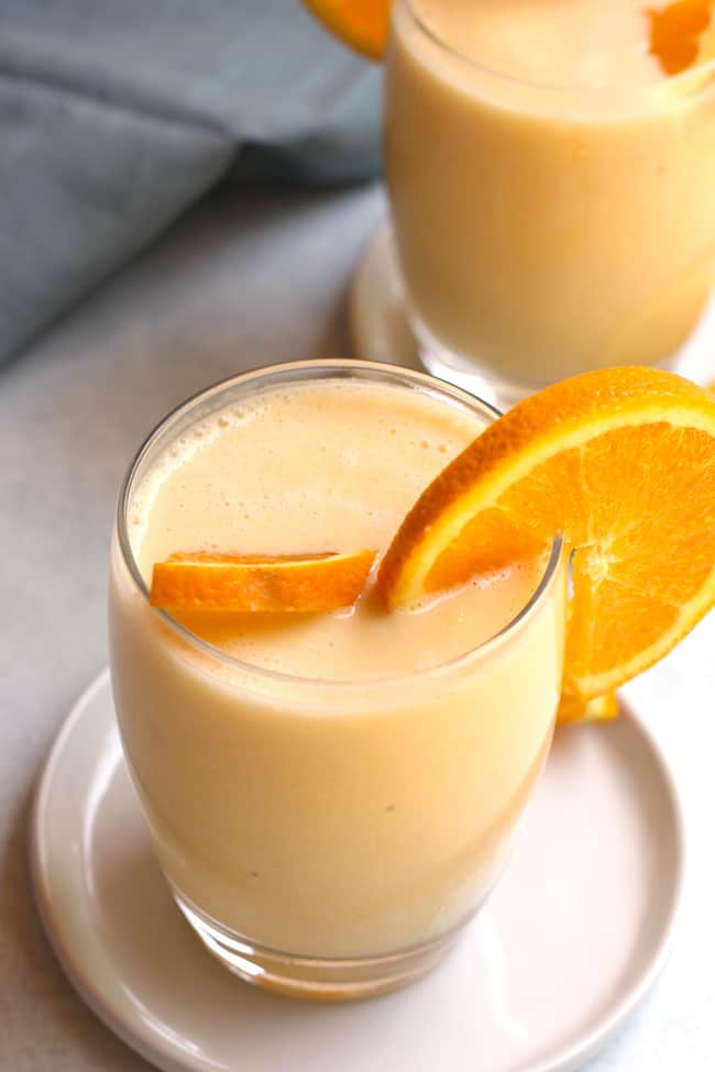 Side shot of two glasses of Orange Julius Smoothie, with orange slices, on small white plates.