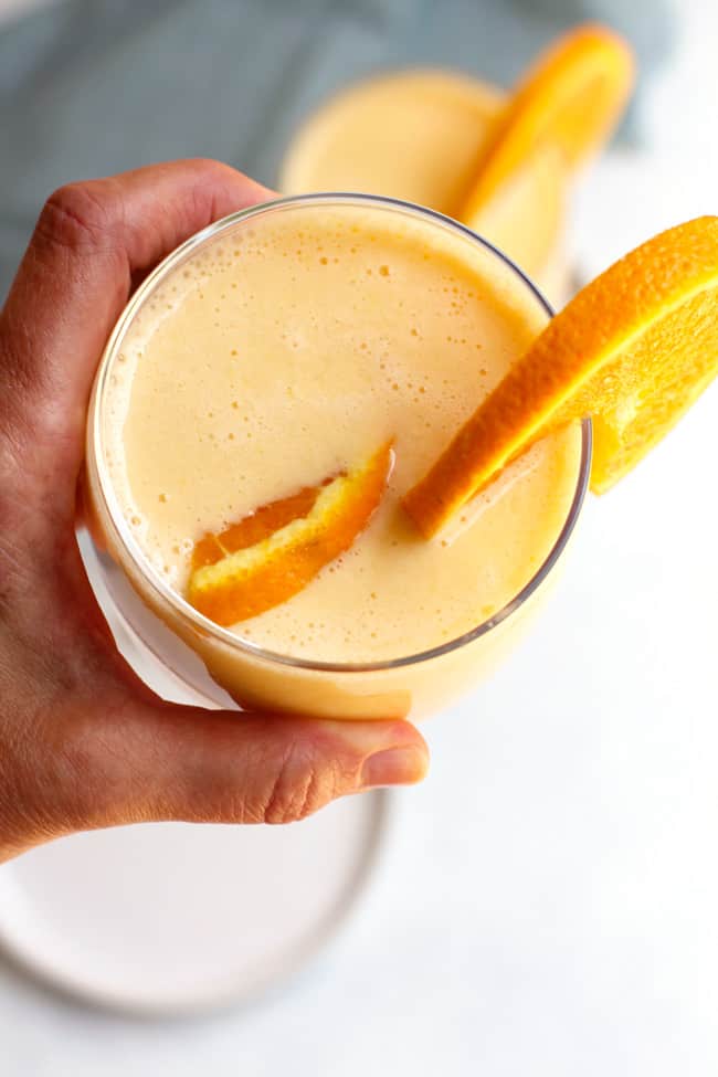 Overhead shot of a hand holding a glass of Orange Julius Smoothie, with a white plate underneath and orange wedges.