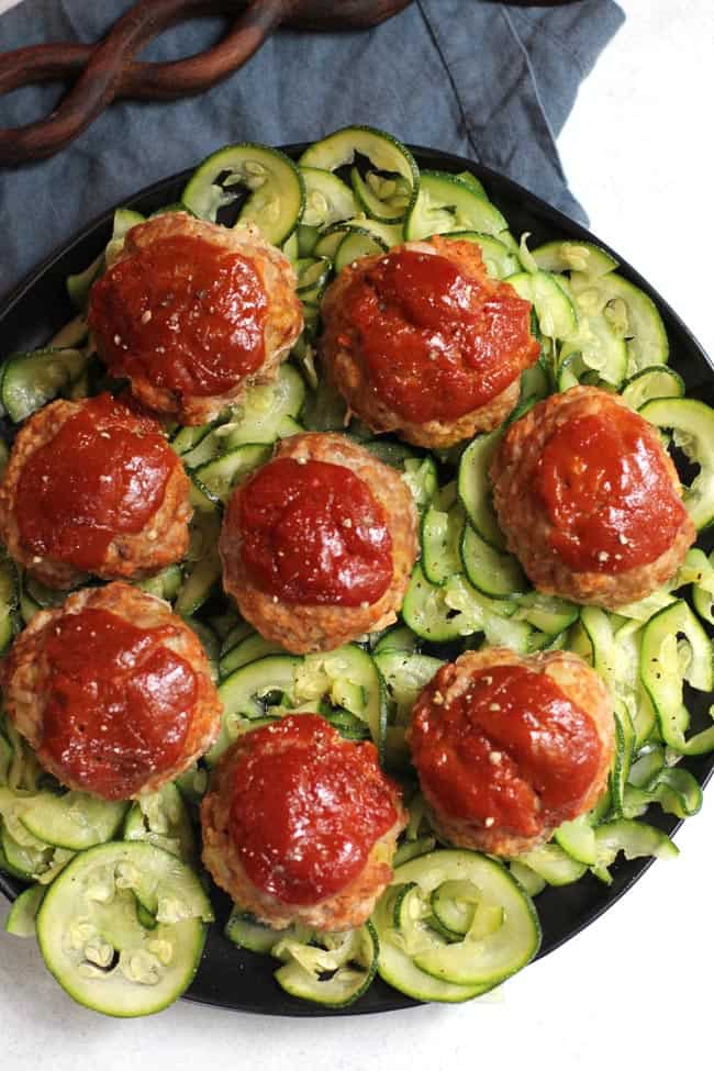 Overhead shot of a black plate of zucchini noodles topped with 8 turkey meatloaf muffins.