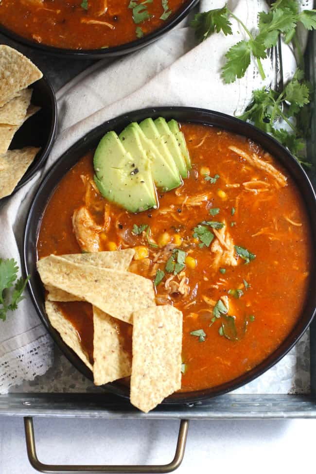 Overhead shot of one black bowl filled with chicken tortilla soup, with chips and avocado, on a gray tray.