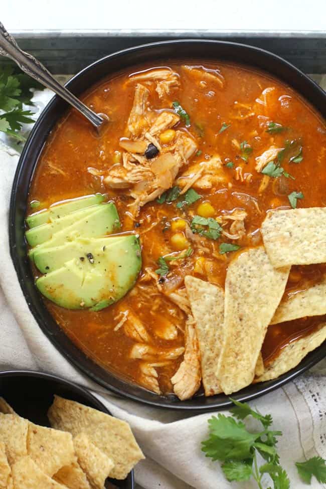 A black bowl full of chicken tortilla soup, with chips and avocado slices.