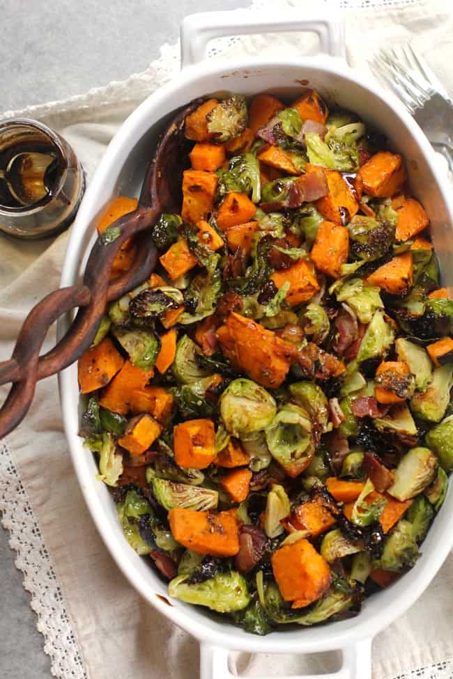 Overhead shot of a oblong white casserole dish of balsamic glazed Brussels sprouts and sweet potatoes, with a wooden spoon.