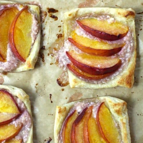 Overhead shot of a sheet pan of baked puff pastry peach tartlets, on tan parchment paper.