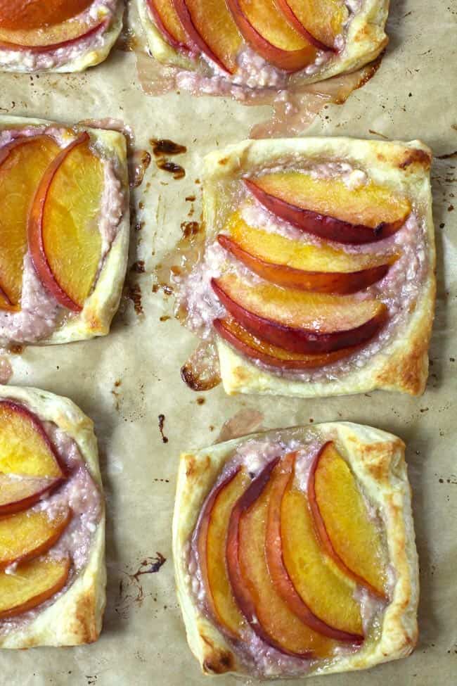 Overhead shot of a sheet pan of baked puff pastry peach tartlets, on tan parchment paper.