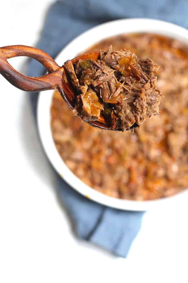 Overhead shot of a wooden spoon filled with shredded beef overtop a white platter of shredded beef, on a blue napkin.