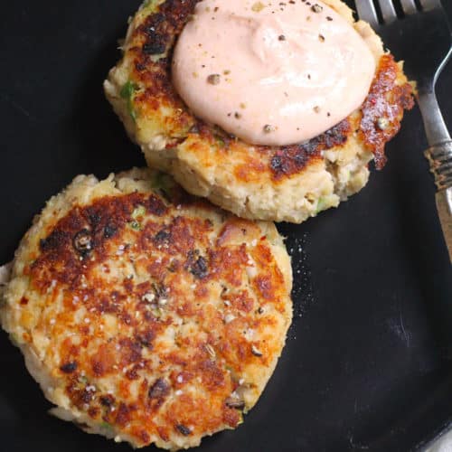 Overhead shot of two tuna patties, on a black plate, one with some sriracha dip on top.