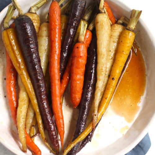 Overhead shot of a round white dish filled with whole multi-colored roasted carrots, on a blue napkin.