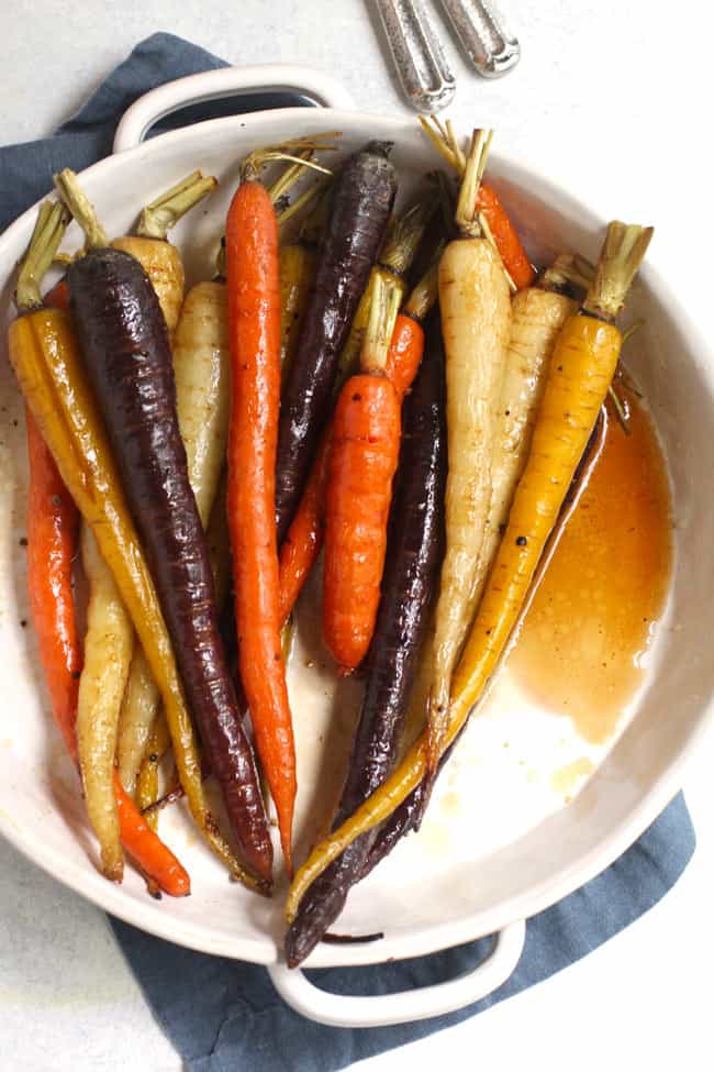 Overhead shot of a round white dish filled with whole multi-colored roasted carrots, on a blue napkin.