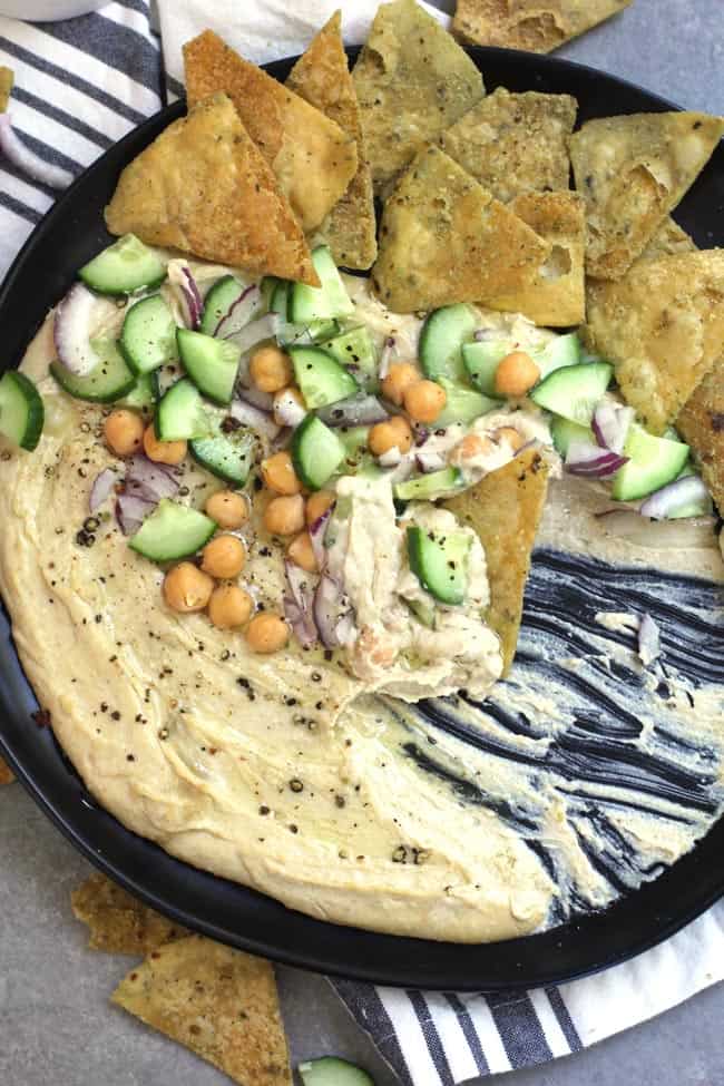 Close-up shot of a partially eaten hummus spread on a black plate, with veggies and pita chips.