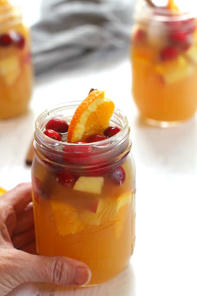 My hand on a jar of apple cider sangria, topped with fruit, with other jars in the background.