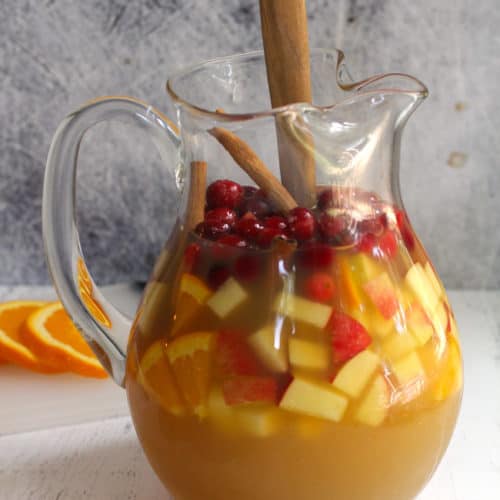 Side shot of a glass pitcher of apple cider sangria filled with fruit, and a wooden spoon inside.