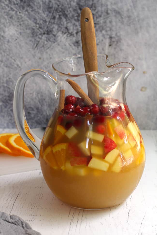 Side shot of a glass pitcher of apple cider sangria filled with fruit, and a wooden spoon inside.