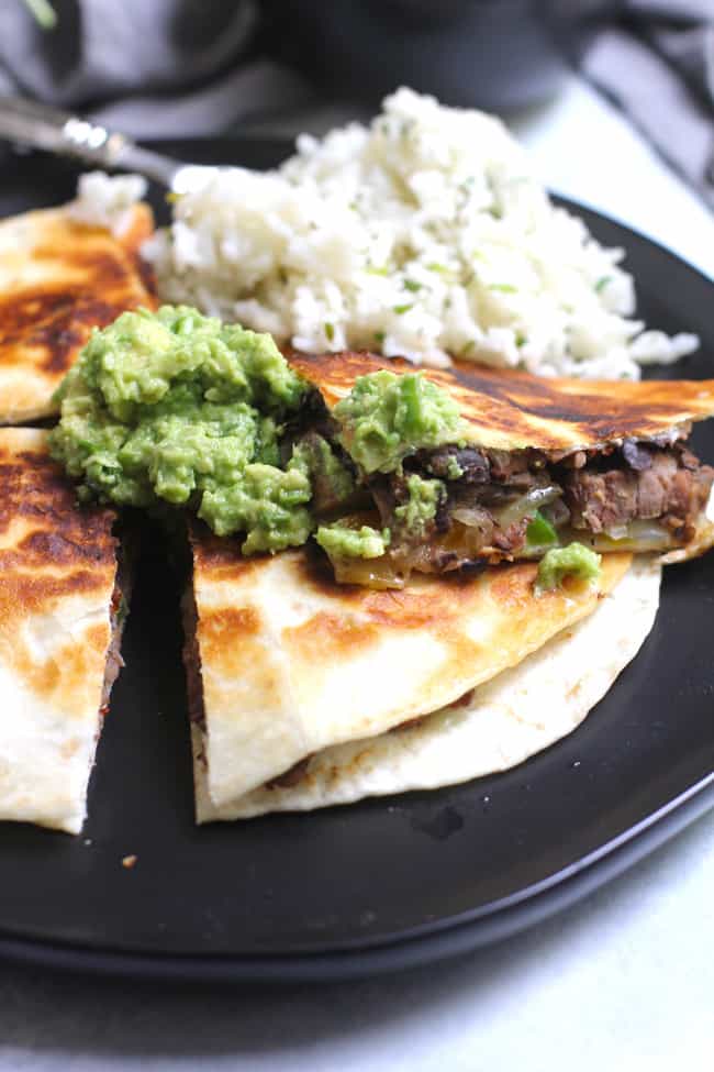 Side shot of a black plate of steak fajita quesadillas, with a scoop of guac and a side of rice.