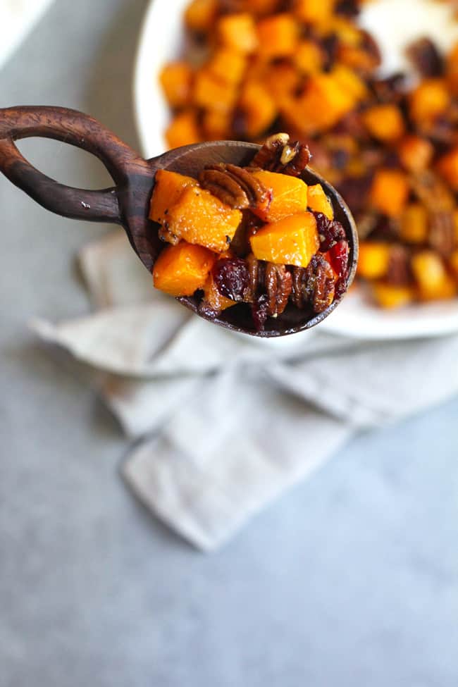 Overhead shot of a wooden spoon of roasted squash with pecans and cranberries, with a dish underneath.