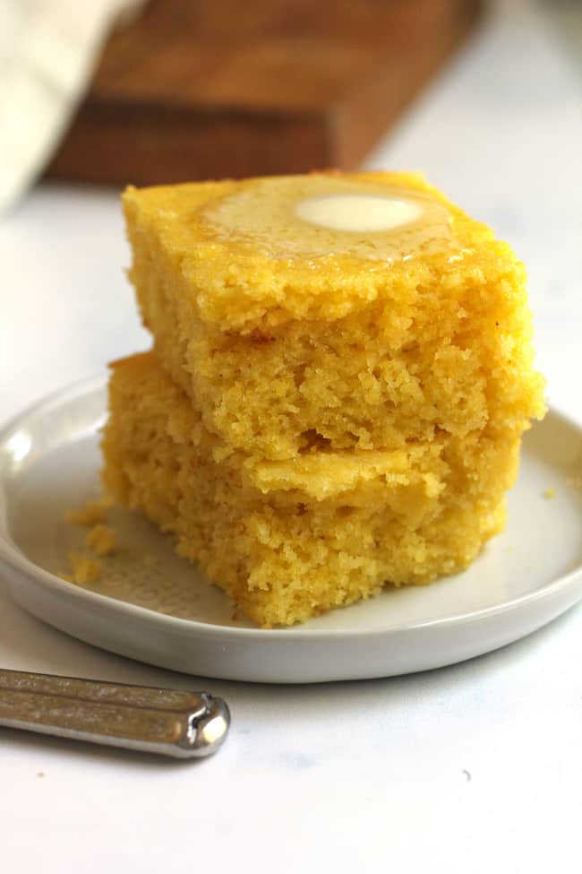 Side shot of two stacked pieces of cornbread on a white plate, with a dollop of melting butter on top.