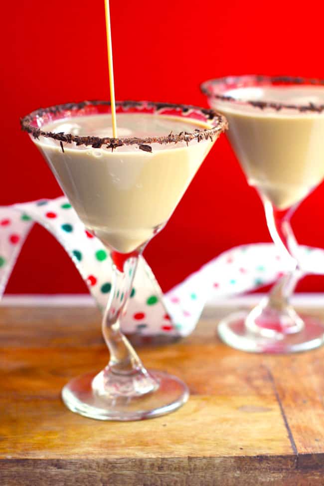 Side shot of two chocolate martinis, with chocolate shavings lining the rims, and a drizzle of martini being poured into one, all with a red background.